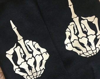 May include: A pair of black ankle socks featuring a white graphic print of a skeletal hand making a middle finger gesture. The design is centered on the top of each sock, showcasing the detailed bone structure of the hand and fingers in a high-contrast white ink against the solid black fabric background.