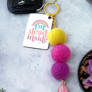 May include: A keychain with a white rectangular charm that says "I'm dead inside" with a rainbow above the text. The keychain has three felt balls, one yellow, one pink, and one purple, and a multi-colored tassel.