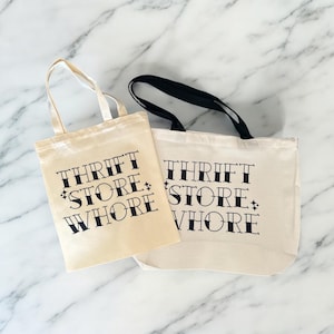 May include: Two white canvas tote bags with black handles. The bags have the text "Thrift Store Whore" printed on them in a black, stylized font.