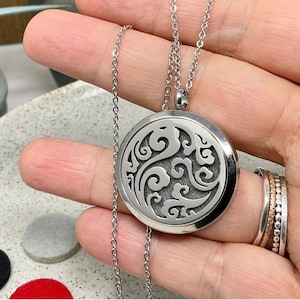 May include: A silver-coloured, round essential oil diffuser necklace with an intricate, swirling design. The pendant hangs from a delicate chain. The necklace is held in a hand, with rings on the fingers. Small, round felt pads in black, grey, and red are visible.