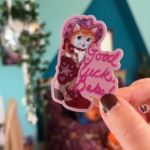 May include: A pink and red glitter sticker featuring a cartoon cat wearing a cowboy hat and boots. The sticker reads "Good Luck, Bebe!"