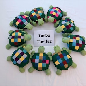 May include: Eight plush turtle toys arranged in a circle around a white rectangular card with the text Turbo Turtles printed in black. Each turtle has a light green head and limbs with a dark green shell featuring a colorful grid pattern.