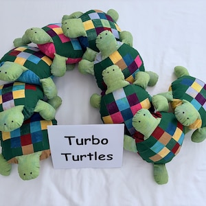 May include: A collection of plush turtle toys with light green heads and limbs, featuring shells made of a multicolored patchwork grid pattern in shades of blue, yellow, red, and green. A white sign in the center displays the text Turbo Turtles.