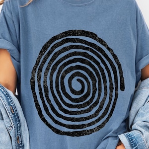 May include: A blue t-shirt featuring a large, distressed black spiral design. The shirt has short sleeves and a relaxed fit. A denim jacket is draped over the shoulders. The overall style is casual and comfortable.