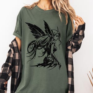 May include: Olive green t-shirt featuring a black illustration of a fairy with detailed wings and long flowing hair. The fairy is depicted in a seated pose, with a distressed, vintage aesthetic. The shirt has a relaxed fit.
