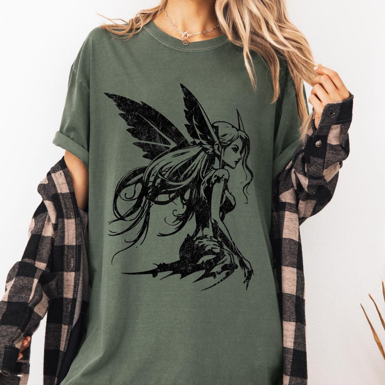 May include: Olive green t-shirt featuring a black illustration of a fairy with detailed wings and long flowing hair. The fairy is depicted in a seated pose, with a distressed, vintage aesthetic. The shirt has a relaxed fit.