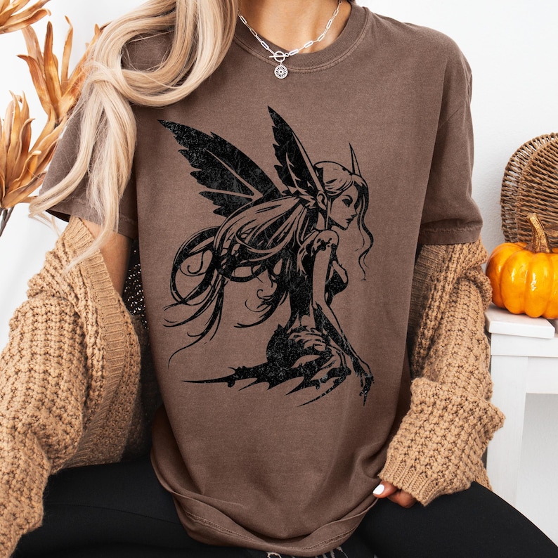 May include: Brown t-shirt featuring a black graphic of a fairy with detailed wings and long hair. The fairy is in a kneeling position. The shirt has a crew neck and short sleeves. The model is wearing a tan cardigan and black pants. A silver necklace is visible.