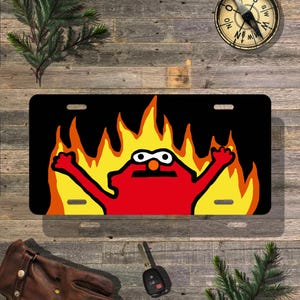 May include: Black license plate featuring a cartoon character in red with a black mustache and white eyes, set against a backdrop of yellow and orange flames. The plate has two mounting holes.