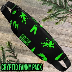 May include: Black fanny pack with a green and black print. The print features the words "Cryptid Research" and various cryptids, including a winged creature, a creature with a long neck, and a creature with a long tail. The fanny pack has a black adjustable strap.