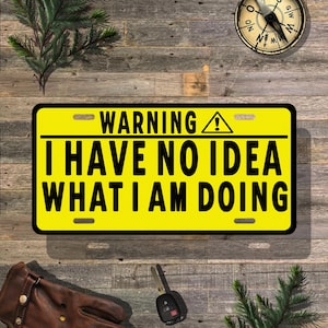 May include: A yellow license plate with black text that reads "WARNING I HAVE NO IDEA WHAT I AM DOING." The plate has a black border and a warning symbol. The background is a wooden surface.