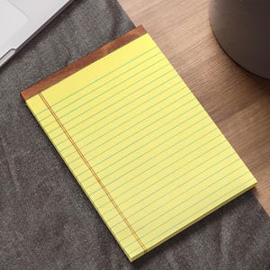 May include: A yellow lined notepad with a brown wooden cover. The notepad is on a gray surface.