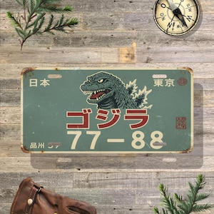 May include: A vintage-style metal license plate with a teal background and a Godzilla graphic. The plate features Japanese text and the numbers "77-88". The top of the plate has the Japanese word for "Japan" and "Tokyo".