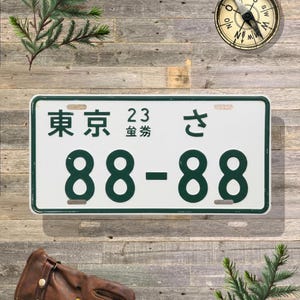 May include: Rectangular white license plate with green trim and black numbers and Japanese characters. The plate reads "東京" above "88-88". A compass and leather glove are in the background.