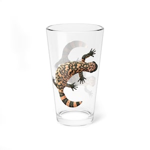 May include: A clear pint glass featuring a Gila monster design. The lizard is orange, black, and tan, with a striped tail. The glass is transparent, and the design appears to be printed on the glass. The glass is empty and sits on a white surface.