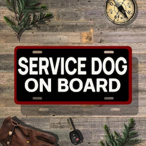May include: A rectangular black sign with the text "SERVICE DOG ON BOARD" in bold white letters, framed by a red border. The sign is on a wooden surface, with a compass, keys, and a leather glove visible.
