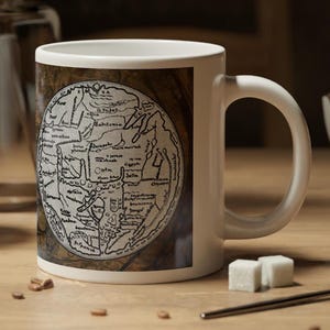 May include: White ceramic mug featuring a detailed, antique map design. The map is circular with intricate lines and text, set against a brown background. The mug has a curved handle and is placed on a wooden surface.