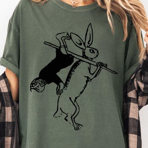 May include: Olive green t-shirt featuring a black illustration of a person and a rabbit holding a stick. The person is upside down, appearing to be riding the rabbit. The design is a whimsical, vintage-style graphic.
