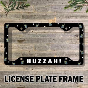 May include: Black license plate frame with the word "Huzzah!" in white. The frame is decorated with cartoon frogs in capes and stars. The words "License Plate Frame" are at the bottom.
