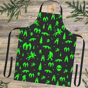 May include: Black apron with a green pattern of aliens, Bigfoot, and other mythical creatures. The pattern is repeated throughout the apron.