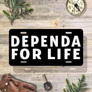 May include: Black license plate with the words "DEPENDA FOR LIFE" in bold white letters. The plate has four mounting holes. The background includes a compass, a car key, a leather glove, and sprigs of greenery.
