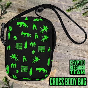 May include: Black crossbody bag with a green print of various cryptids, including a ghost, a winged creature, a tree, and an alien. The bag has the text "CRYPTID RESEARCH TEAM" repeated on the front.