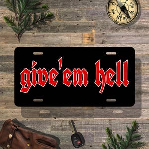 May include: Black license plate with the phrase "give 'em hell" in red gothic lettering. The plate has four mounting holes and is displayed on a wooden surface with a compass, keys, and a leather glove.