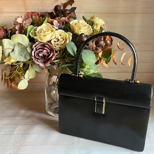 May include: A structured black handbag with a smooth finish, a single arched top handle, and a rectangular body. The bag features a central metallic clasp closure and gold-toned hardware accents where the handle attaches to the main frame.
