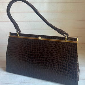 Peut inclure: Un sac à main vintage marron avec une texture semblable à celle d'un crocodile. Le sac rectangulaire est doté d'un fermoir et d'une garniture dorés, ainsi que d'une poignée incurvée. Le sac est conçu pour être porté à la main.