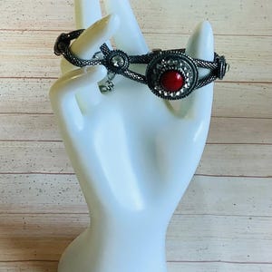 Può includere: Bracciale argentato con pietra centrale rossa e pietre chiare di contorno. Il bracciale è esposto su un espositore bianco a forma di mano, su sfondo in legno chiaro. Chiusura con catena.