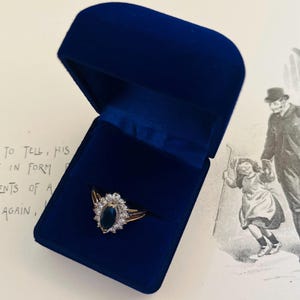 May include: A gold ring with a dark blue oval gemstone, encircled by smaller white stones, presented in an open, dark blue velvet ring box. The box is set against a background with a vintage illustration.