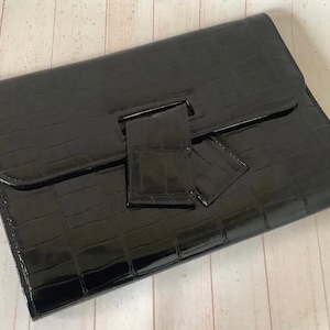 以下が含まれることがあります： クロコダイル型押しで光沢のある仕上げの黒いクラッチバッグ。ユニークな折り返し式クロージャーを採用しています。