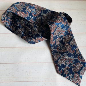 Könnte beinhalten: Eine marineblaue Krawatte mit einem Paisley-Muster in Rot, Weiß und Gold. Die Krawatte besteht aus einem seidigen Material und wird auf einer hellen Holzoberfläche präsentiert. Das Paisley-Muster ist ein klassisches Design.