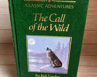 Op de afbeelding: Een groen hardcover boek met de titel "The Call of the Wild" van Jack London. De cover heeft gouden letters en een illustratie van een wolf die naar de maan huilt. Onderdeel van de "Classic Adventures" serie.