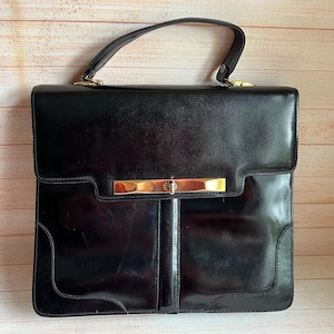 Könnte beinhalten: Eine schwarze Lederhandtasche mit goldenem Verschluss und Griff. Die rechteckige Tasche hat ein strukturiertes Design mit Klappenverschluss und oberem Griff. Das glänzende Finish und die goldfarbene Hardware verleihen der Tasche einen klassischen, eleganten Look.