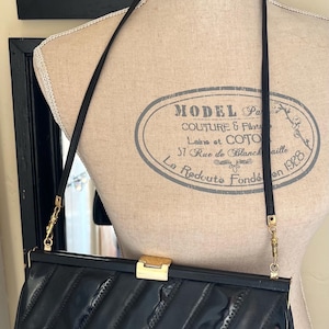 Könnte beinhalten: Schwarze rechteckige Handtasche mit diagonalem Steppmuster, goldfarbenem Verschluss und langem schwarzem Riemen. Die Tasche wird auf einer Schaufensterpuppe präsentiert. Die Schaufensterpuppe hat ein Vintage-Design mit dem Text "MODEL Pa COUTURE & Pilat".