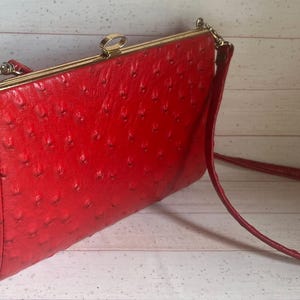 May include: A bright red rectangular handbag with a textured surface featuring small, evenly spaced indentations. The bag has a gold-toned metal frame, a top clasp closure, and a thin matching red shoulder strap attached to the sides.