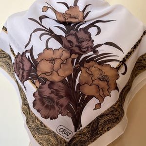 May include: White cotton scarf with a brown and beige floral print. The scarf has a paisley border and the word "CROSS" printed in the center.