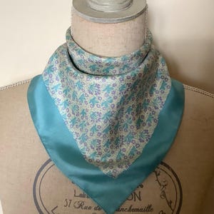 May include: A teal blue silk scarf with a floral pattern in shades of purple and green. The scarf has a teal blue border.
