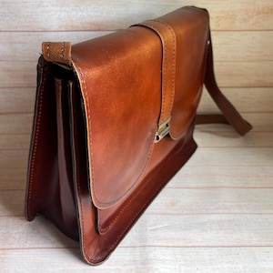 May include: A structured brown leather satchel featuring a smooth finish, a fold-over flap with a vertical strap detail, and a metallic silver-toned clasp closure. The bag includes a side gusset design and an attached shoulder strap for carrying.