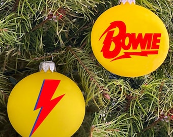 May include: Two yellow Christmas ornaments with red and blue designs. One ornament features the word "BOWIE" in red letters with a lightning bolt design. The other ornament features a red lightning bolt design.