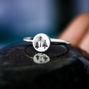 May include: A silver ring with a circular design featuring three stylized pine trees. The ring is resting on a smooth, dark gray stone.