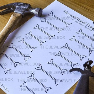 Könnte beinhalten: Ein weißes Blatt Papier mit einer Meerjungfrauen-Schwanz-Schablone darauf gedruckt. Das Papier ist von Werkzeugen umgeben, darunter ein Hammer und ein Werkzeug aus Metall mit einem Griff.