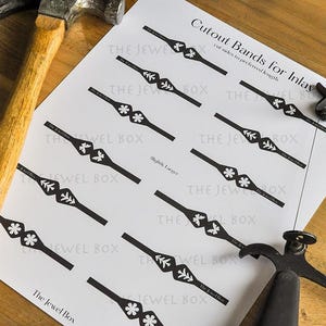 May include: A sheet of paper with black cutout band designs for inlay, with text that reads "Cutout Bands for Inlay". A hammer and a tool are on the wooden surface. The Jewel Box is printed on the paper.