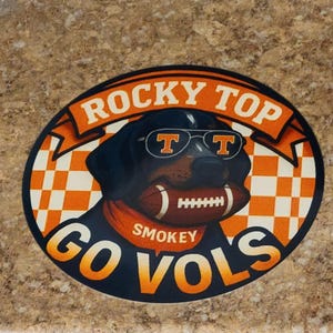 May include: Oval sticker featuring a black dog wearing sunglasses with the letters "T" and holding a football. The sticker has the words "Rocky Top" and "Go Vols" in orange and white.