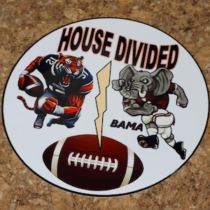 May include: A circular decorative item featuring a tiger and an elephant, each holding a football. The text "HOUSE DIVIDED" is at the top, with "BAMA" below the elephant. A football graphic is at the bottom.