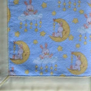 May include: A blue baby blanket with a yellow border. The blanket features a repeating pattern of white bunnies sitting on yellow crescent moons and clouds with yellow stars hanging from them.