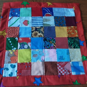 May include: A patchwork quilt with a red border featuring colorful star and square patterns. The quilt's center is composed of various fabric squares with floral, fruit, and geometric designs. The quilt measures approximately 18 inches by 18 inches.