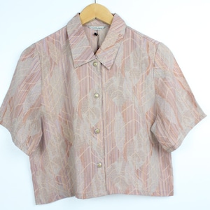 May include: A short-sleeved button-down shirt in a neutral color palette of beige, peach, and light brown. The shirt features a subtle leaf pattern and is hanging on a wooden hanger against a white background.