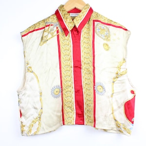 May include: Cream-colored sleeveless shirt with a red and gold pattern. The shirt features vertical red stripes down the front and a gold chain and floral design. The collar is red and the shirt is hanging on a wooden hanger.