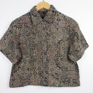 May include: A short-sleeve button-up shirt featuring a dense, intricate tapestry-style pattern in shades of black, brown, tan, and muted purple. The garment has a standard collar, a front button placket, and a straight hemline with a textured finish.